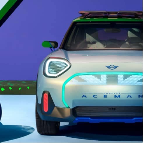 Front view of a MINI Concept Aceman parked on a light blue surface with an illustrative dark sky and trees in the background.