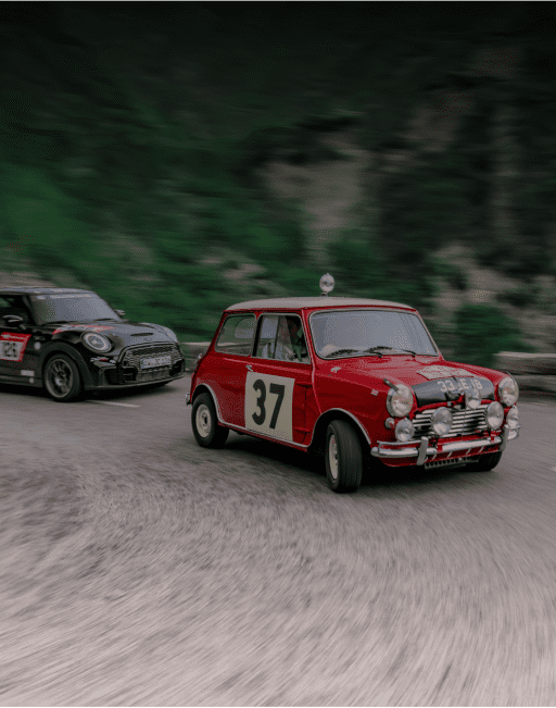A red vintage MINI Cooper turning around a bend with a second MINI following close behind it with blurred cliffs covered with greenery in the background.