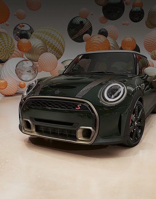 Three-quarters front view of the MINI Cooper SE Electric Hardtop Resolute Edition in Rebel Green, parked in a CGI world with different colored balloons floating in the background.