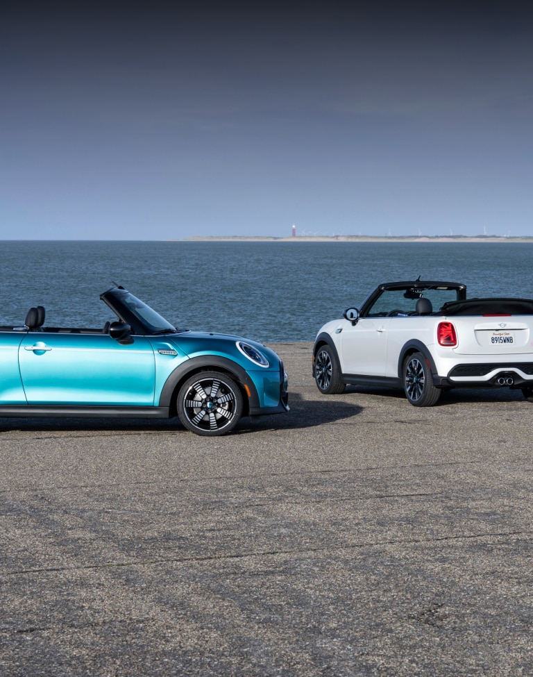 Side view of a MINI Seaside Edition in the Caribbean Aqua metallic body color parked on a beach next to another MINI Seaside Edition in the Nanuq White colorway positioned in a three-quarters back view with a body of water and clear skies in the background.