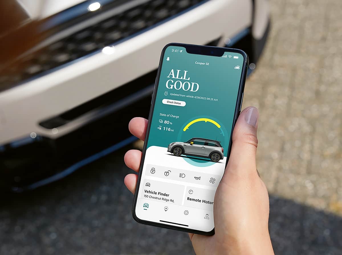 Hand holding a smartphone with MINI App on the screen and front of a white MINI vehicle in the background.