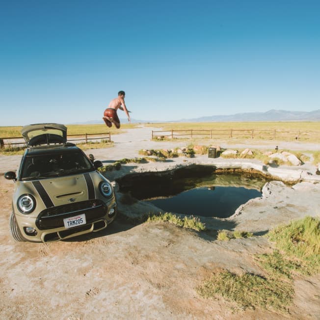 Green MINI with black bonnet stripes parked near a hot spring with a shirtless man in the air along with a mountain range and clear skies in the background.