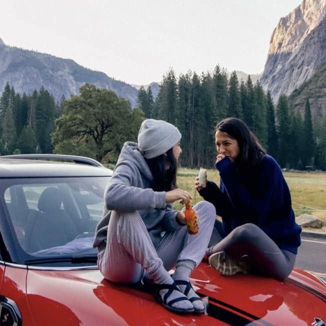 Two women sitting and laughing on the hood of a red MINI vehicle with evergreen trees and mountains in the background.