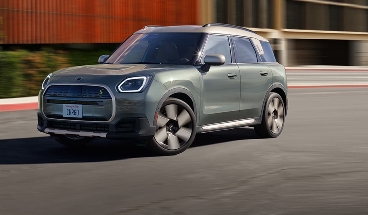 Three-quarters front view of a 2025 MINI Countryman SE ALL4 driving on a street in an urban setting with its shadow underneath the vehicle and its surroundings blurred out.