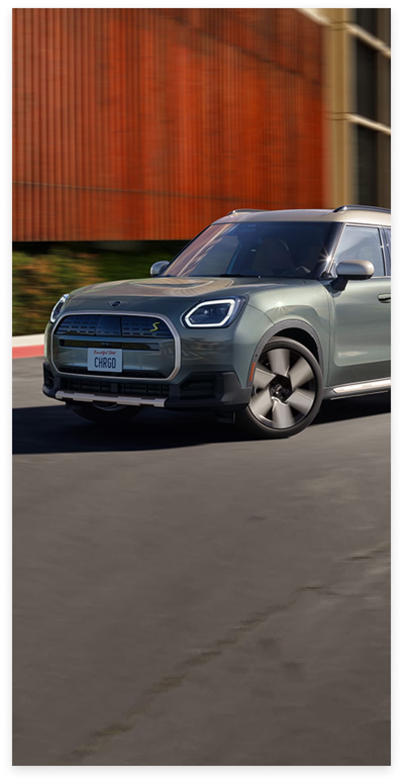 Three-quarters front view of a 2025 MINI Countryman SE ALL4 driving on a street in an urban setting with its shadow underneath the vehicle and its surroundings blurred out.