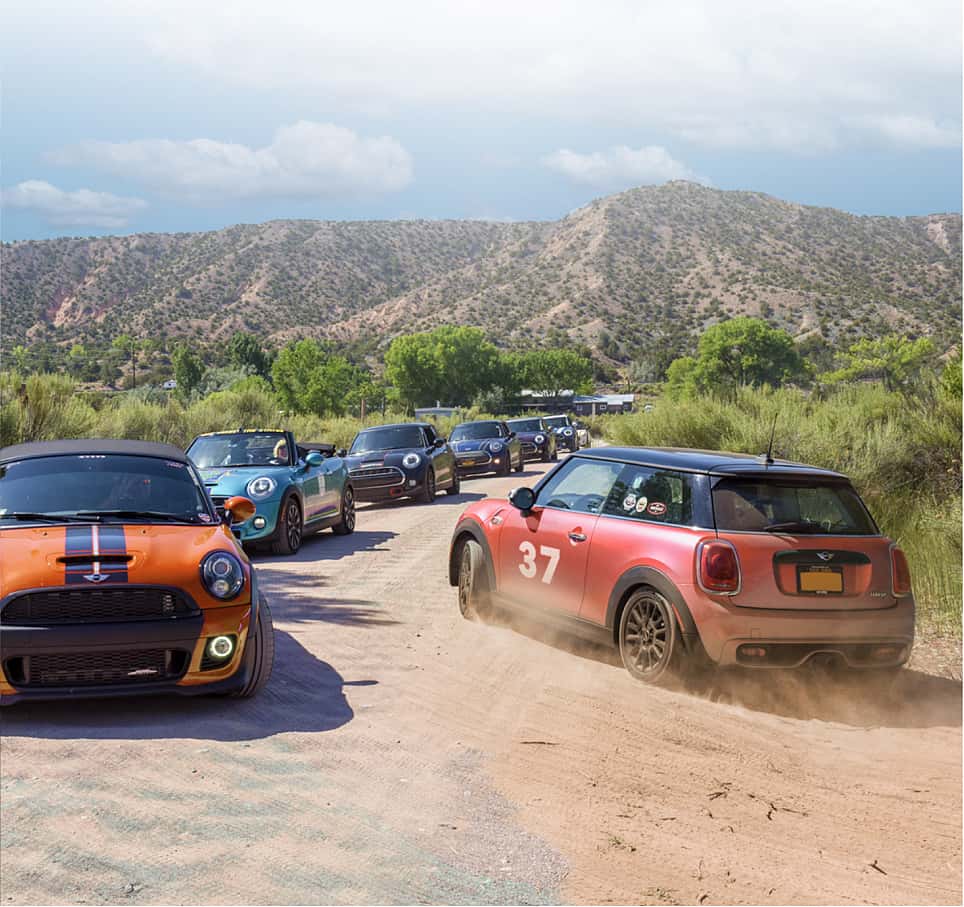 Several MINI vehicles in different colors driving on a dirt path with plants and trees surrounding them along with mountains and cloudy skies in the background.