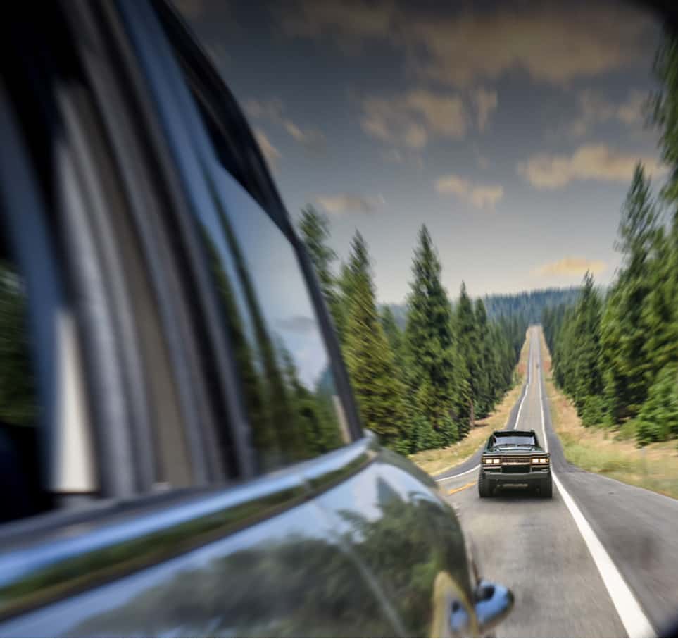 Fisheye view of a vehicle behind another vehicle driving on an open road with evergreen trees and clear skies in the background.