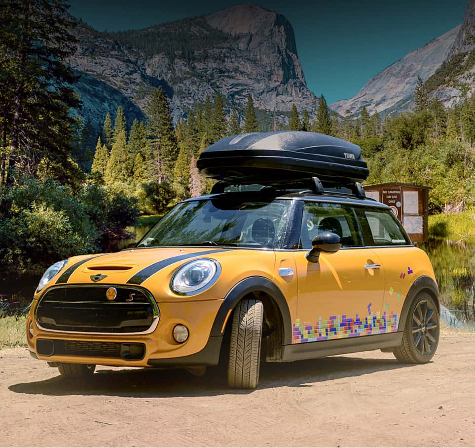 Orange MINI with black bonnet stripes and colored tile decals on the side, parked on a dirt surface with evergreen trees and mountains in the background.