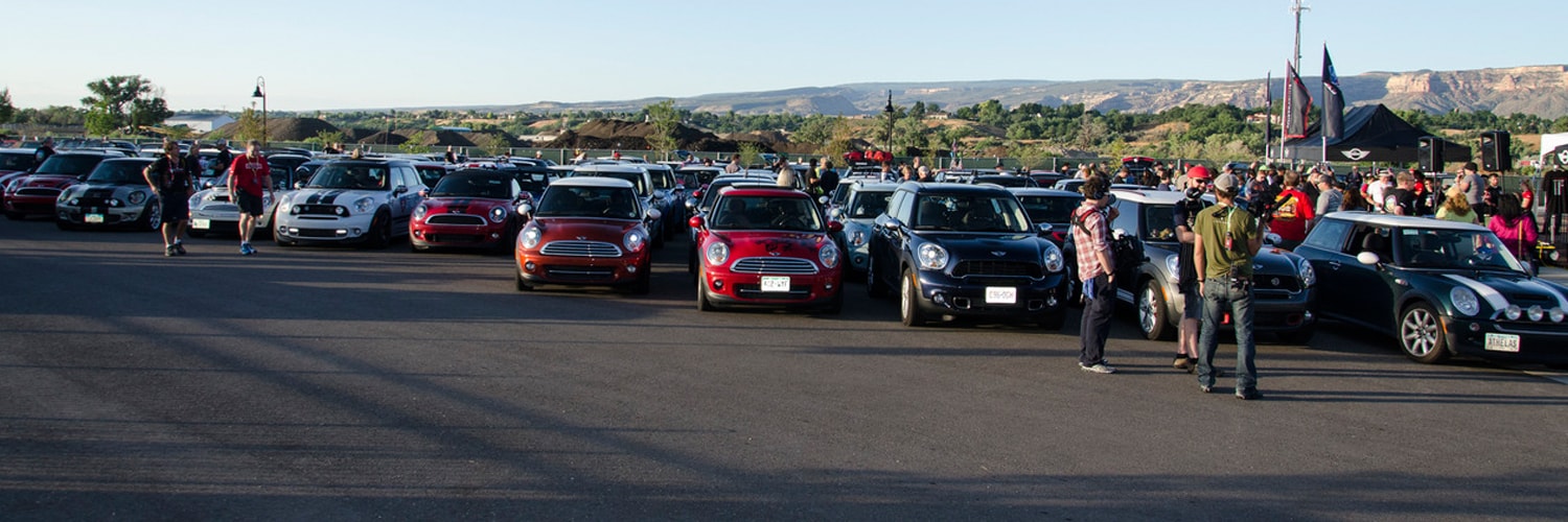 Landscape view of several MINI vehicles of different colors parked in a parking lot, with people walking nearby or standing around in both the foreground and background, on a sunny day with clear skies, trees, mountains, and a MINI tent in the background.