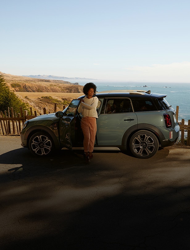 Side view of a four-doored MINI vehicle parked on pavement, with a woman in a white sweater exiting the driver’s seat along with a coastal view in the background on a cloudless day.