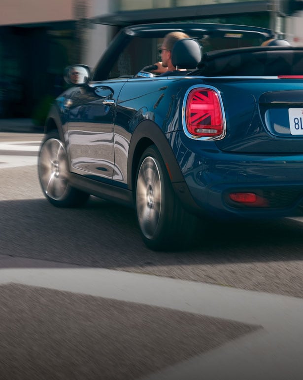 Three-quarters back view of a blue MINI Convertible, with a specific focus on the back-left Union Jack taillight, driving on a street with its surroundings blurred out.