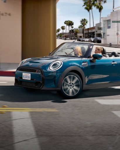 Three-quarters front view of a blue MINI Convertible rounding a street corner in an urban setting with palm trees and clear blue skies in the background.