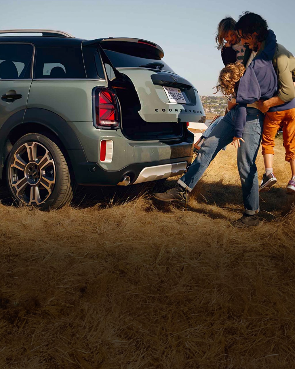 Back view of a dark green MINI Countryman with a family standing behind the vehicle and using the Automatic Tailgate function while the trunk is swing open.