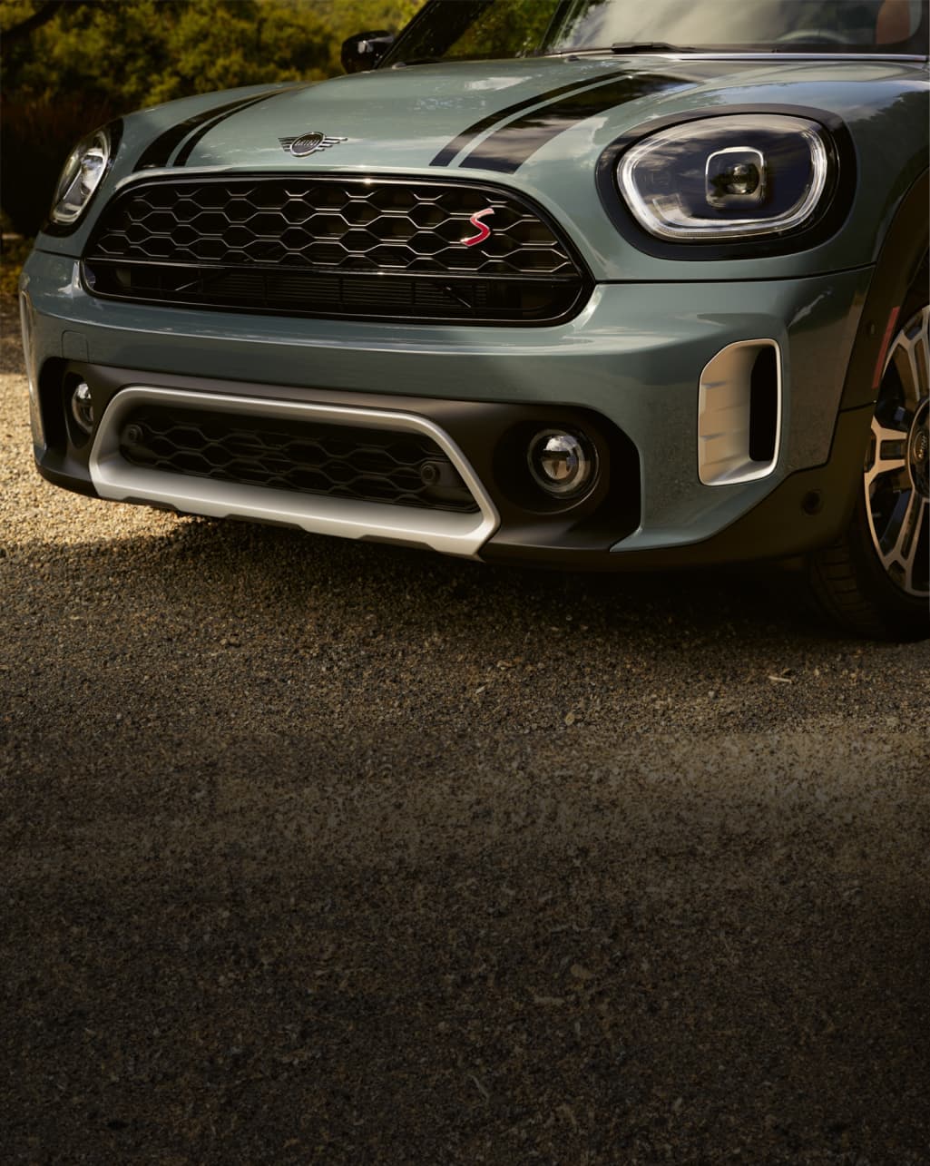 Three-quarters view of the front part of a dark green MINI Countryman parked on a pavement surface, including the patterned grille and LED headlights.
