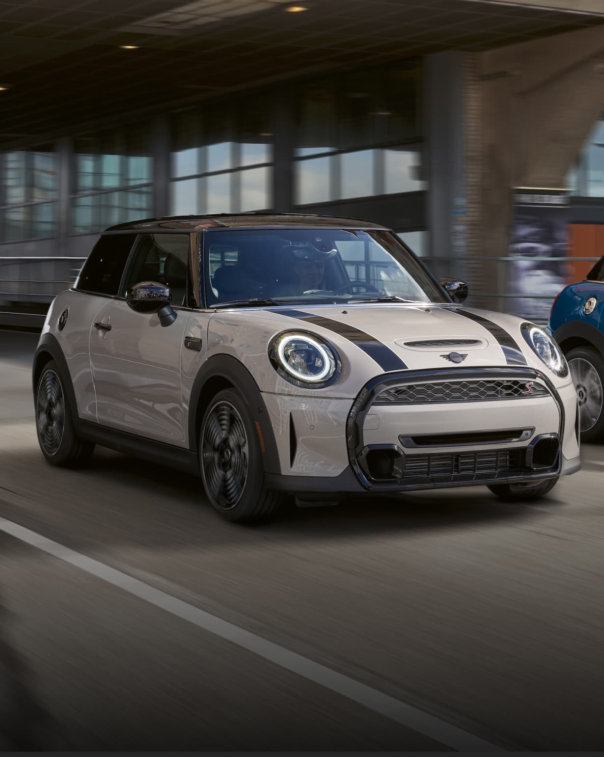 Front view of a grey MINI Hardtop 2 Door driving on a street in an urban setting with its surroundings blurred out.