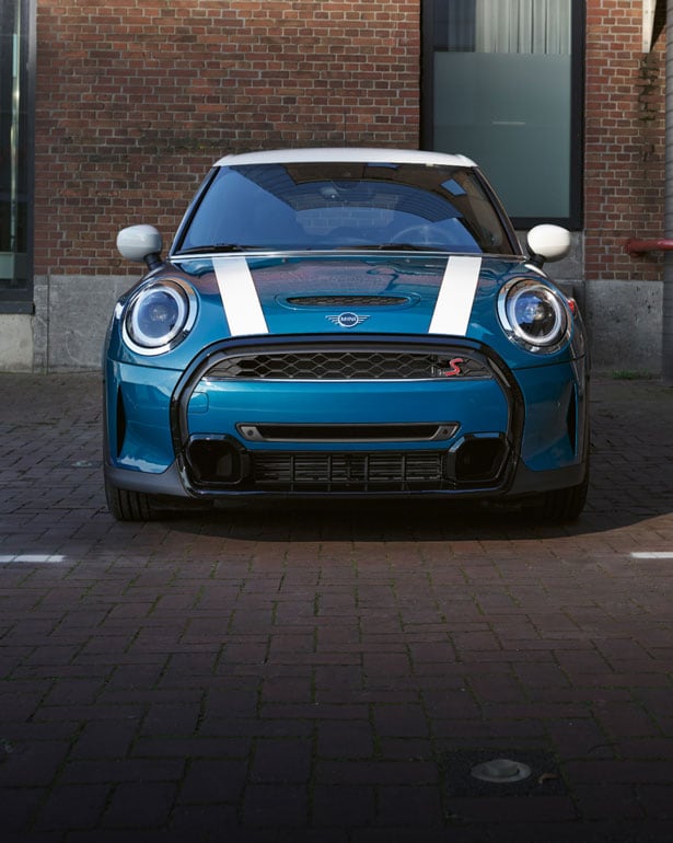 Front view of a blue MINI Hardtop 4 Door with a white roof and white bonnet stripes, parked in a parking space with a brick wall and window in the background.