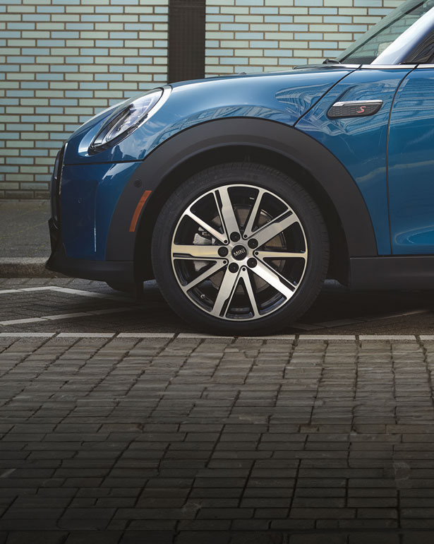 Side view of a blue MINI Hardtop 4 Door parked in a cobblestone parking space with a brick wall in the background.